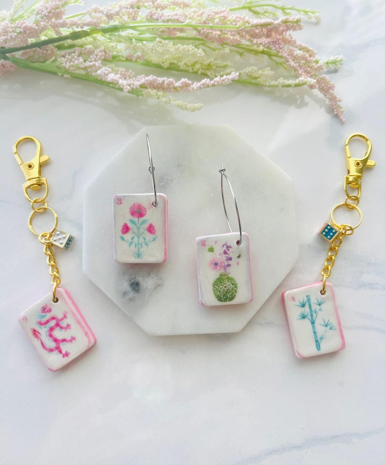 May include: Keychains and earrings featuring rectangular charms with floral and oriental designs. The charms have a white background with pink borders. The keychains have gold-colored clasps and dice charms. The earrings have silver hoops.