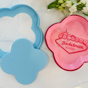 Puede incluir: Un cortador de galletas azul claro y un sello de galletas azul claro a juego están junto a un plato rosa con un borde rojo. El plato tiene las palabras "Welcome to Bachelorette Weekend" en un diseño retro.