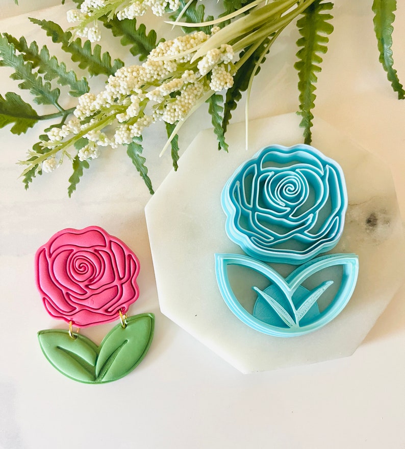 Op de afbeelding: Een roze roosvormige oorbel met groene bladeren en goudkleurige connectoren. Daarnaast een blauwe roosvormige koekjesvorm met bladvormige uitsteekvormen. De items liggen op een wit oppervlak met een takje witte bloemen.