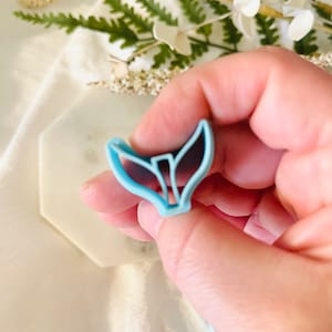 May include: A light blue plastic mermaid tail cookie cutter. The cutter is shaped like a mermaid tail with a fin on top and a forked tail on the bottom.