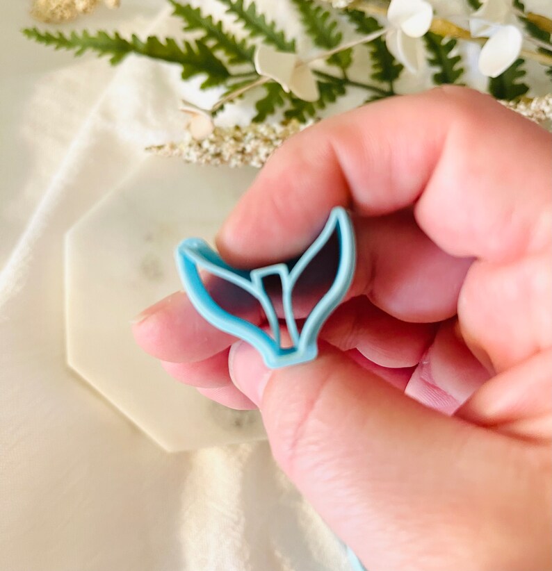 May include: A light blue plastic mermaid tail cookie cutter. The cutter is shaped like a mermaid tail with a fin on top and a forked tail on the bottom.