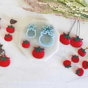 Könnte beinhalten: Eine Sammlung handgefertigter Accessoires zum Thema Tomate, darunter Ohrringe, ein Schlüsselanhänger und Ausstecher. Die Ohrringe zeigen rote Tomaten mit grünen Stielen. Der Schlüsselanhänger hat einen einzelnen Tomatenanhänger. Ausstecher sind hellblau.