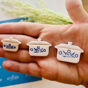 May include: Three miniature white ceramic casserole dishes with blue floral designs. The dishes are arranged in a row, varying in size. Each has a lid and a handle. The design is reminiscent of vintage kitchenware.