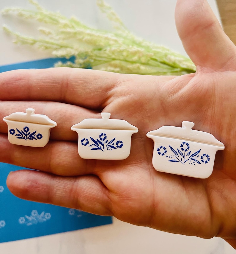 May include: Three miniature white ceramic casserole dishes with blue floral designs. The dishes are arranged in a row, varying in size. Each has a lid and a handle. The design is reminiscent of vintage kitchenware.