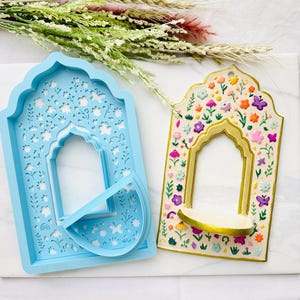 Può includere: Uno stampo in silicone azzurro a forma di finestra ad arco con intricati ritagli di fiori e farfalle, accanto a una mensola da parete ad arco color crema decorata con fiori e farfalle dipinti a colori vivaci con bordo dorato.