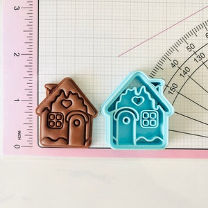 May include: Two gingerbread house cookie cutters, one brown and one light blue. The brown cutter is slightly larger than the blue cutter. Both cutters have a heart-shaped window, a door, and a chimney.