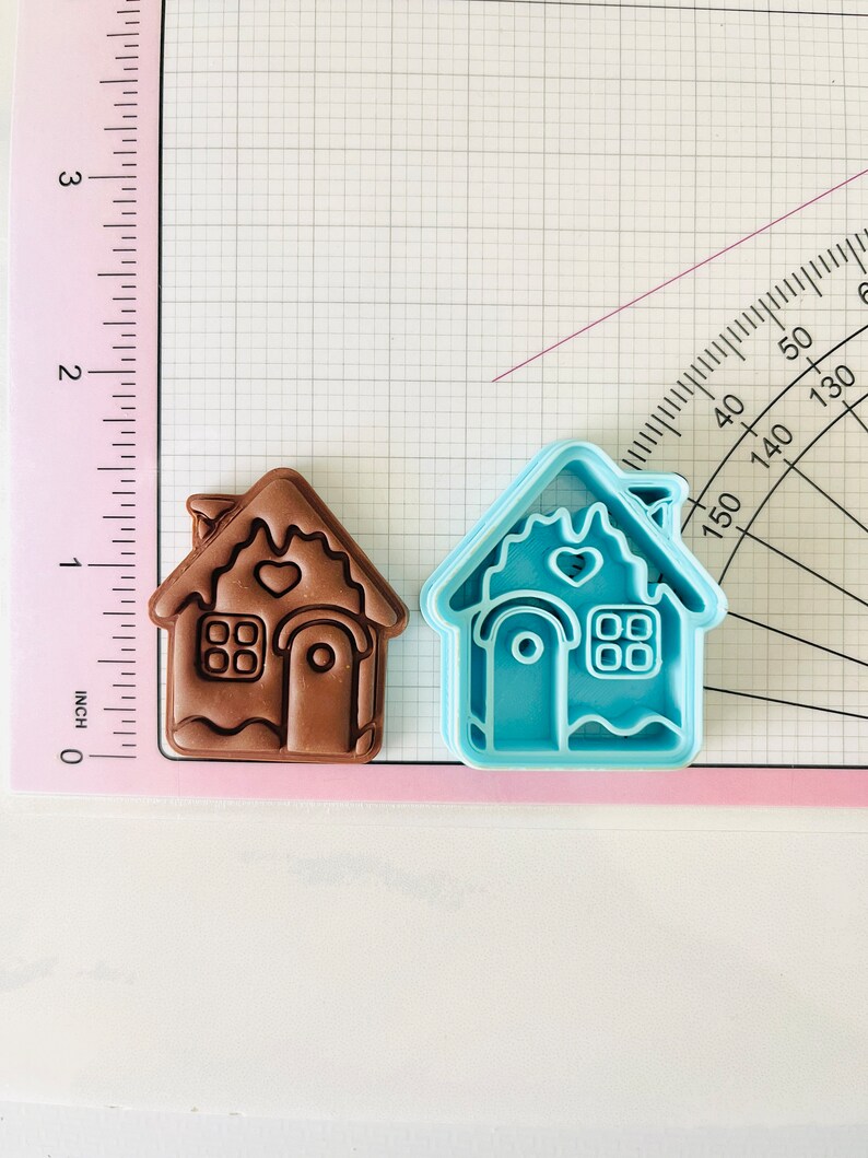 May include: Two gingerbread house cookie cutters, one brown and one light blue. The brown cutter is slightly larger than the blue cutter. Both cutters have a heart-shaped window, a door, and a chimney.