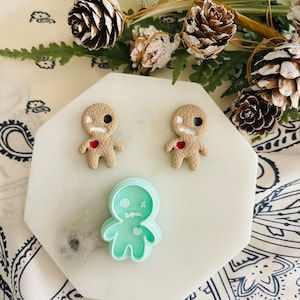 May include: Two brown gingerbread man-shaped polymer clay earrings with red hearts and black eyes. A light blue gingerbread man-shaped cookie cutter is also shown.