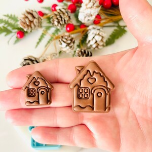 May include: Two brown gingerbread house shaped cookie cutters, one larger than the other. Both have a heart-shaped window and a door with a rounded top.