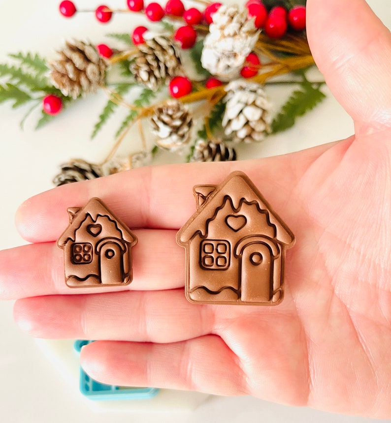 May include: Two brown gingerbread house shaped cookie cutters, one larger than the other. Both have a heart-shaped window and a door with a rounded top.
