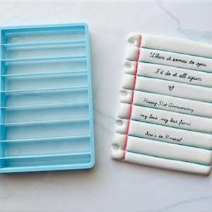 Op de afbeelding: Een lichtblauwe plastic koekjessnijder naast een witte koek die is versierd om op een notitieblok te lijken. De notitieblokkoek heeft tekst in zwarte inkt, waaronder "When it comes to you" en "Happy 31st Anniversary."