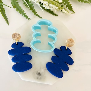 Może przedstawiać: Para niebieskich kolczyków z polimerowej gliny z złotym akcentem. Kolczyki mają kształt serii połączonych owali. Kolczyki są prezentowane na białym marmurowym podłożu z niebieską plastikową formą.