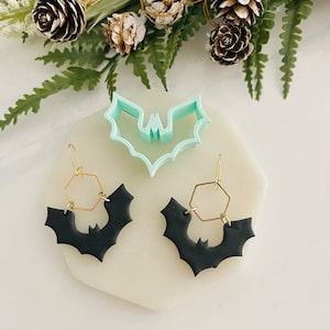 May include: A pair of black bat-shaped earrings with gold hexagonal hoops. The earrings are displayed on a white surface with a green plant in the background.