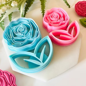 Op de afbeelding: Een verzameling roosvormige koekjessnijders in verschillende kleuren. De uitstekers omvatten een lichtblauwe roos, een roze roos en een roze bladuitsteker. De uitstekers zijn gemaakt van plastic en zijn gerangschikt op een wit oppervlak.