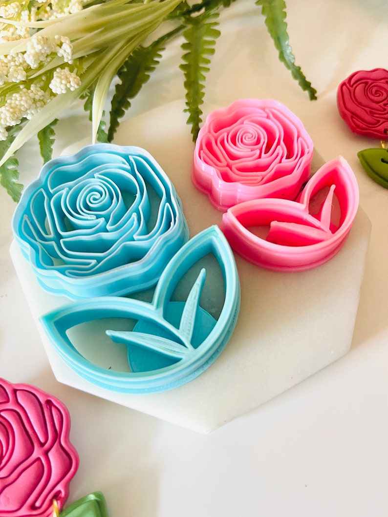 Op de afbeelding: Een verzameling roosvormige koekjessnijders in verschillende kleuren. De uitstekers omvatten een lichtblauwe roos, een roze roos en een roze bladuitsteker. De uitstekers zijn gemaakt van plastic en zijn gerangschikt op een wit oppervlak.