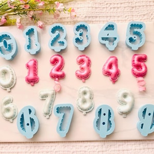 May include: A collection of light blue plastic number cookie cutters from zero to nine, alongside pink and white molded number shapes that resemble balloon designs, arranged on a flat surface for baking or crafting purposes.