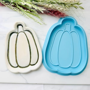 May include: A white ceramic pumpkin-shaped dish with dark green outlines alongside a bright blue plastic pumpkin-shaped cookie cutter. The dish features a smooth surface with a raised stem and vertical ridges, while the cutter has a matching outline.