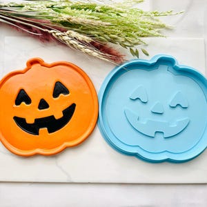 May include: Two pumpkin-shaped items, one orange with a black carved face and one light blue with a matching carved face design. The orange item has a smooth finish, while the light blue item appears to be a mold or a flat-surfaced object.