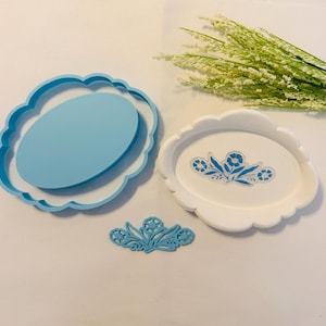 May include: A light blue oval cookie cutter with a scalloped edge, a white oval tray with a scalloped edge featuring a blue floral design in the center, and a separate light blue floral-shaped stamp piece for creating detailed impressions.