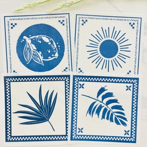 Pode incluir: Quatro bases de copos quadradas brancas com desenhos azuis. Cada base de copo apresenta um design diferente: um lim&atilde;o, um sol, uma folha de palmeira e uma folha de samambaia. As bases de copos t&ecirc;m uma borda azul com um padr&atilde;o decorativo.