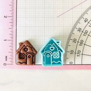 May include: Two gingerbread house cookie cutters, one brown and one blue. The brown cutter has a heart and window details. The blue cutter has a heart, window, and door details.