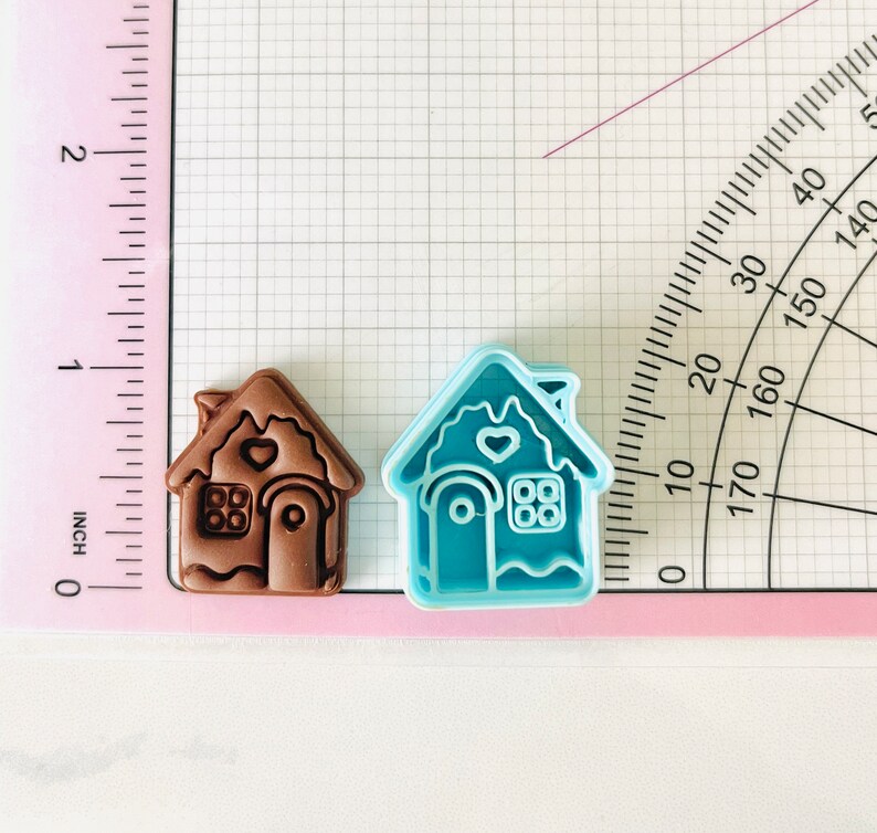 May include: Two gingerbread house cookie cutters, one brown and one blue. The brown cutter has a heart and window details. The blue cutter has a heart, window, and door details.