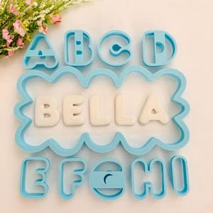 Könnte beinhalten: Ein Set aus hellblauen Buchstaben- und Namens-Ausstechern aus Kunststoff. Der Name "BELLA" wird in weißen Fondantbuchstaben in einem wellenförmigen Rahmen angezeigt. Einzelne Buchstaben-Ausstecher A, B, C, D, E, F, G, H und I sind ebenfalls enthalten.