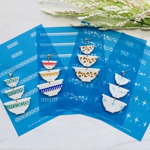 Op de afbeelding: Handgemaakte witte en gouden oorbellen met een komontwerp. De oorbellen zijn gemaakt van gelaagde, patroon keramische stukken. De oorbellen worden gepresenteerd op een blauwe achtergrond met witte patronen.