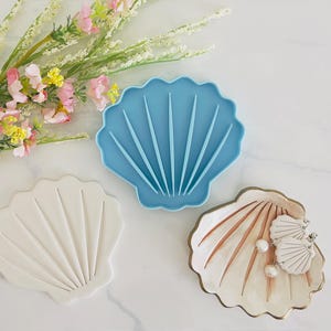 May include: Three seashell-shaped trays in different colours. A light blue tray, a white tray, and a beige tray with gold trim. The beige tray holds two seashell earrings with pearl accents. Decorative floral arrangement in the background.