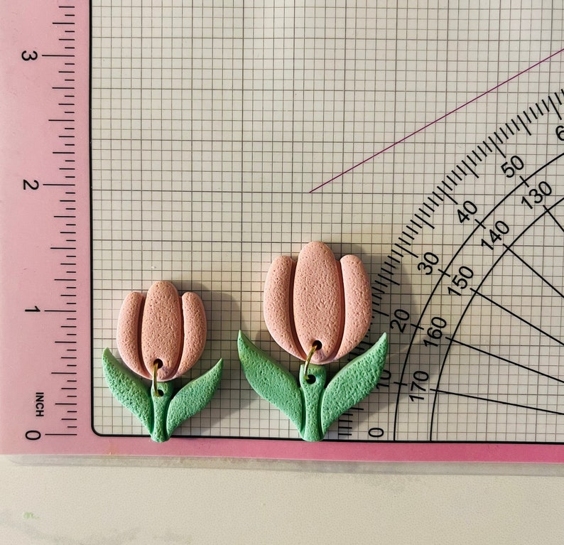 May include: A pair of pink and green polymer clay earrings shaped like tulips. The earrings have gold hooks.