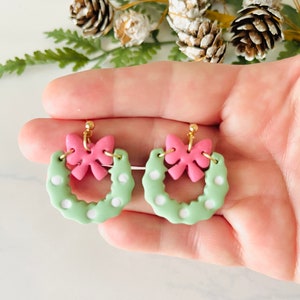 May include: A pair of green and pink Christmas wreath earrings with white polka dots and a pink bow on each wreath.