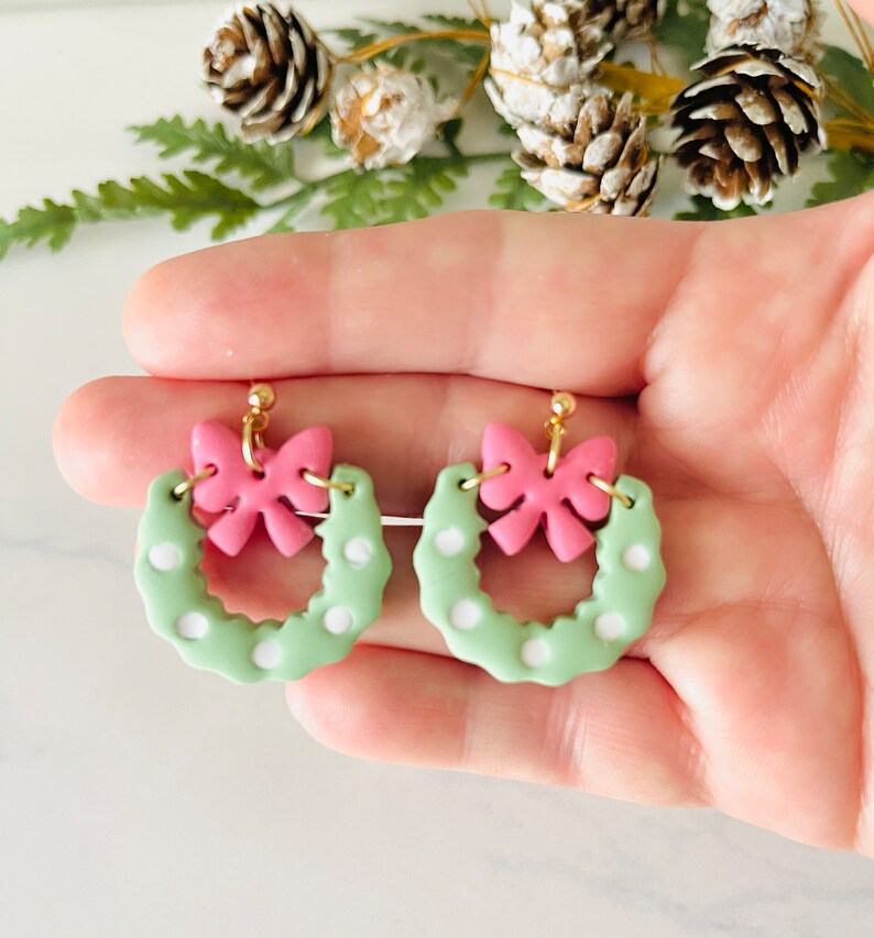May include: A pair of green and pink Christmas wreath earrings with white polka dots and a pink bow on each wreath.