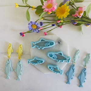 Puede incluir: Pendientes y cortadores de galletas con forma de pez en tonos azules y amarillos. Los pendientes tienen una parte superior con forma de pez y un colgante de pez más grande. Los cortadores de galletas son de varios tamaños. Flores coloridas en el fondo.