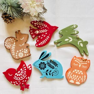 Puede incluir: Conjunto de seis adornos de madera pintados a mano, con forma de ardilla, pájaros, conejo y búho. Cada adorno presenta intrincados diseños florales y de hojas blancos. Los adornos están ensartados con hilo para colgar.