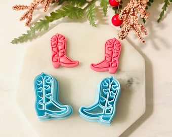 Puede incluir: Cuatro cortadores de galletas con forma de botas de vaquero. Dos son rosas y dos son azules.