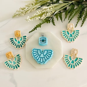 Puede incluir: Cuatro pares de pendientes de cerámica azul y blanca con detalles dorados. Los pendientes tienen forma de abanico y un diseño geométrico. También hay dos moldes de plástico azul para hacer los pendientes.