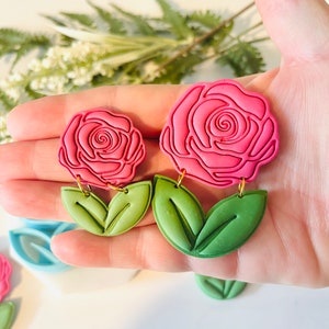 Op de afbeelding: Roze roosvormige oorbellen met groene bladeren. De oorbellen hebben een gedetailleerd rozenontwerp met een goudkleurige haak. De bladeren zijn lichtgroen. Een bloemig accessoire.