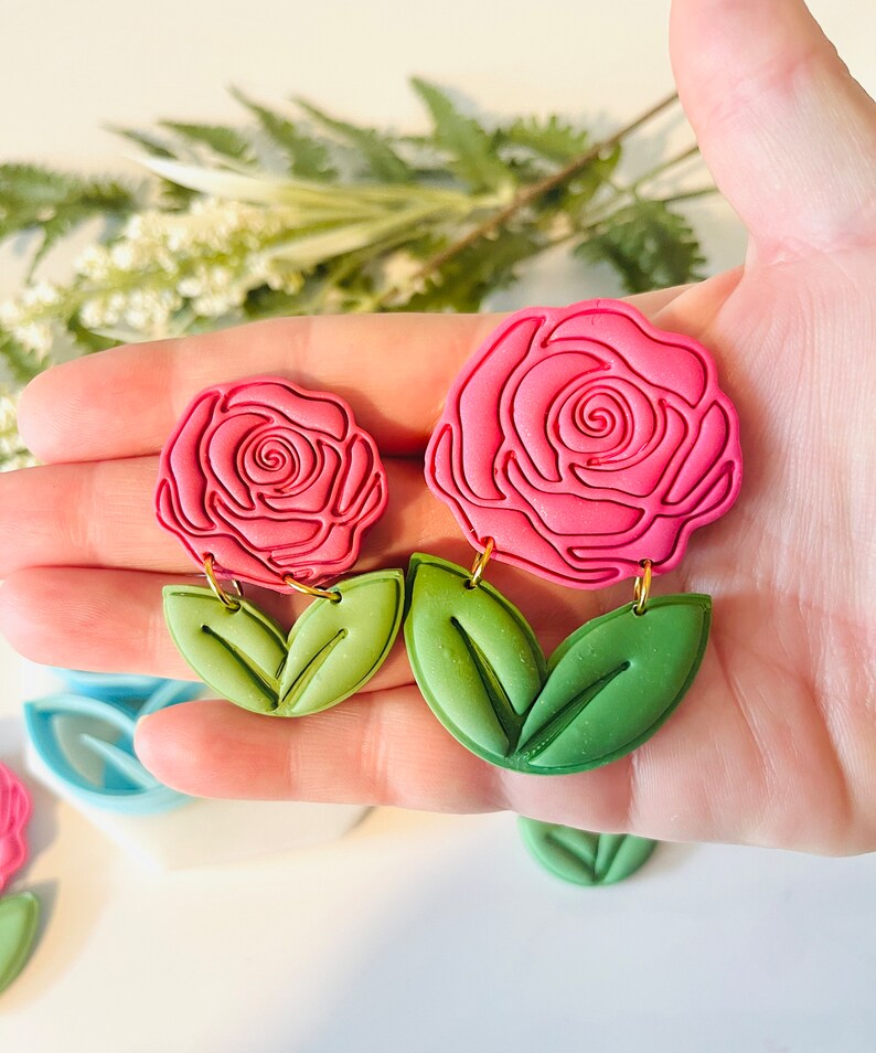 Op de afbeelding: Roze roosvormige oorbellen met groene bladeren. De oorbellen hebben een gedetailleerd rozenontwerp met een goudkleurige haak. De bladeren zijn lichtgroen. Een bloemig accessoire.