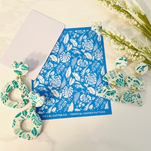 May include: A pair of white and teal blue earrings with a tropical leaf pattern. The earrings are made of polymer clay and have gold-colored hoops. The earrings are displayed on a blue background with a white tropical leaf pattern stencil. The stencil says "So Cal Cutter Co. Tropical Leaves Pattern".