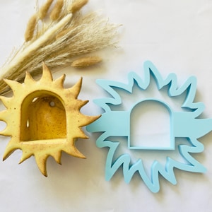 May include: A yellow ceramic sun-shaped wall planter with a speckled finish and a light blue plastic sun-shaped cookie cutter. The ceramic planter features a central arched opening and a small hole for hanging, while the cutter mirrors the sun design.
