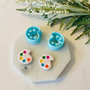 Puede incluir: Dos cortadores de galletas de plástico azul con forma de paletas de pintura y dos decoraciones de galletas pintadas de blanco con puntos de pintura de colores.