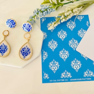 May include: A pair of blue and white porcelain dangle earrings with gold accents. The earrings are displayed on a white surface with a blue and white damask pattern stencil. The stencil has the text "SO CAL CUTTER CO. - ARABESQUE PATTERN".