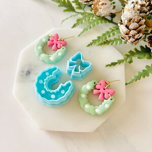 May include: A pair of green and white wreath earrings with pink bows. The earrings are made of polymer clay and have a light blue and white polka dot pattern. The earrings are displayed on a white marble surface.