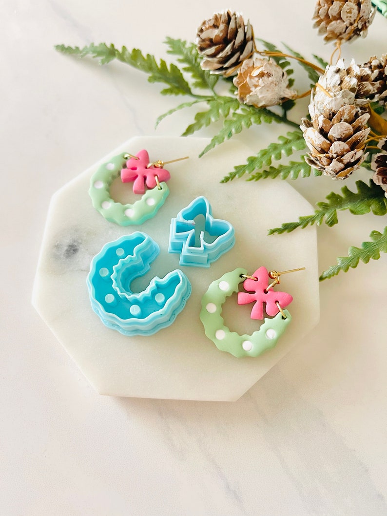 May include: A pair of green and white wreath earrings with pink bows. The earrings are made of polymer clay and have a light blue and white polka dot pattern. The earrings are displayed on a white marble surface.