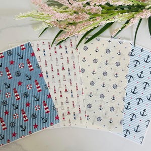 May include: A selection of nautical-themed fabric sheets featuring patterns of lighthouses, anchors, ship wheels, and stars in red, white, and blue. The fabrics are arranged with a sprig of pink flowers in the background.