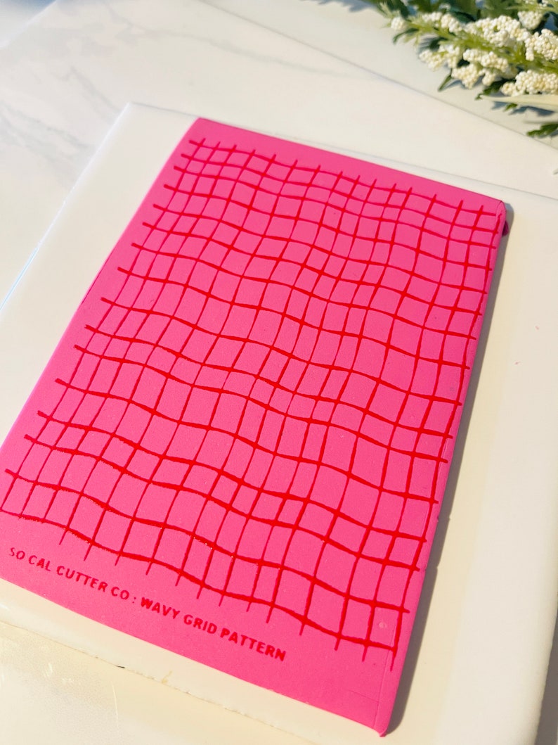 Może przedstawiać: R&oacute;żowa silikonowa mata do pieczenia z falistym wzorem kratki. Mata ma nadrukowany tekst "SO CAL CUTTER CO: WAVY GRID PATTERN" na spodzie.
