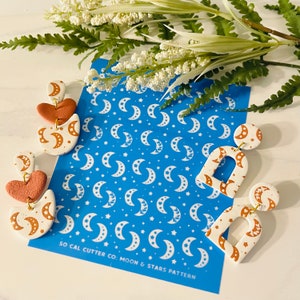Pode incluir: Dois pares de brincos de argila branca e marrom com um padrão de lua e estrelas. Os brincos estão em uma folha de estêncil azul com o texto "So Cal Cutter Co. Moon & Stars Pattern".