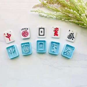May include: A collection of Mahjong tiles in white and light blue. The white tiles feature red and black symbols, including a "JOKER" tile. The light blue tiles have matching symbols. The tiles are arranged on a white surface with green and white floral accents.
