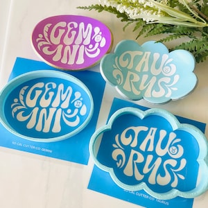 May include: Four colorful Gemini and Taurus themed cookie cutters and trays. The Gemini items are oval-shaped, with a purple tray and a blue cutter. The Taurus items are cloud-shaped, with a light blue tray and a blue cutter.