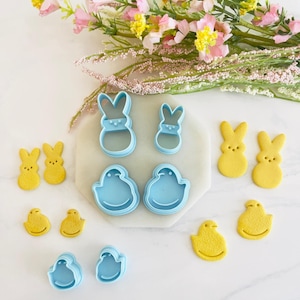 May include: A set of blue plastic cookie cutters in the shapes of bunnies, chicks, and peeps. The cutters are arranged on a white surface with yellow sugar cookies cut out in the same shapes.
